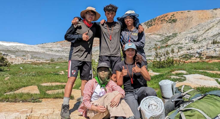 Five people pose for a photo in front of a mountainous ridge. Two of them are giving the camera thumbs up. 